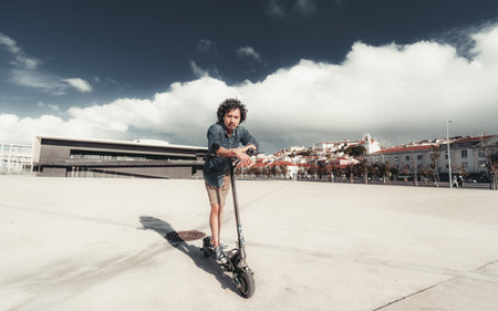 A young asian man with curly hair leans casually on an electric scooter in a spacious, urban setting with modern architecture in the background. Cloudy sky, relaxed pose, warm lightの写真素材