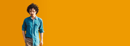 Young asian man with curly hair wearing a blue denim shirt poses against a bright orange background with his hand in his pocket. Vibrant portrait with copy space area on the rightの写真素材