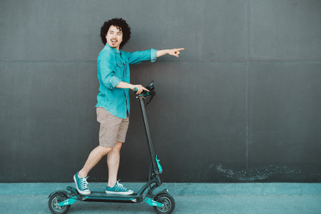 Young man with curly hair riding an e-cooter against a dark wall background. Wearing casual clothes,...