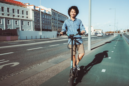 Young man with curly hair riding an electric scooter on a bike path in an urban area. Dressed in a casual denim shirt, shorts, and sneakers, enjoying a sunny day outdoorsの写真素材
