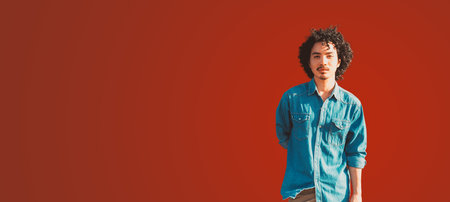 Portrait of a young man with curly hair wearing a denim shirt, standing against a vibrant reddish background. The subject faces the camera with a calm expression, a copy space area on the leftの写真素材
