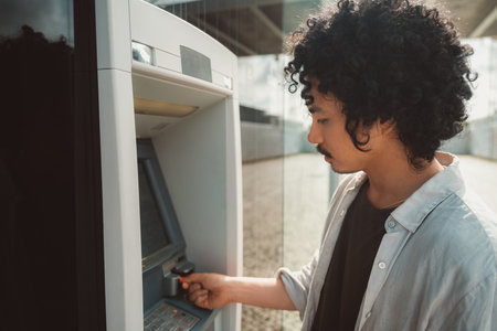 A wide-angle shot of a young man with curly hair and a mustache stands at an outdoor ATM, focused on the screen as he inserts his bank card. Dressed casually in a light shirt and dark t-shirtの写真素材