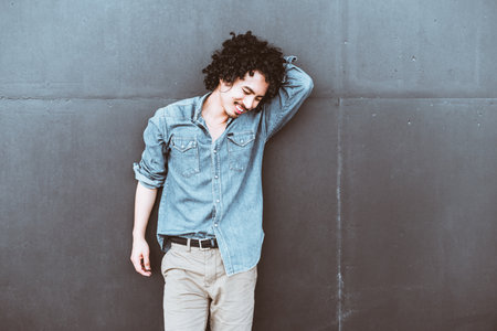 A young man with curly hair wearing a casual denim shirt and light checkered trousers poses against a plain gray wall, smiling and leaning his arm on his headの写真素材