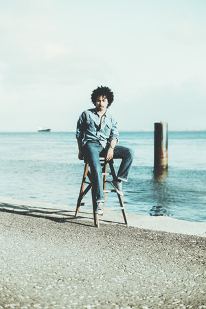 A young mixed man with curly hair sitting confidently on a wooden ladder by a calm waterfront. He wears denim jeans, a denim shirt, and sneakers, exuding a casual styleの写真素材