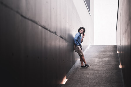 A young biracial man with curly hair in casual attire leans against a modern concrete wall in an urban alleyway, smiling while talking on the phone, with natural light creating depthの写真素材