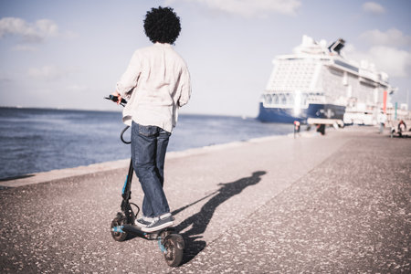 A person with curly hair rides an electric scooter along a waterfront promenade with a large cruise ship docked in the background on a bright, sunny day; a copy space area on the rightの写真素材