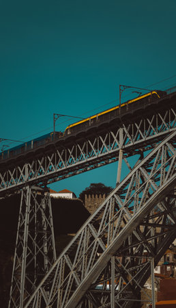 A vibrant yellow tram crosses the upper deck of the Dom Luis I Bridge in Porto, Portugal, with a metro train; an intricate iron structure illuminated by warm sunlight against a clear turquoise skyの写真素材