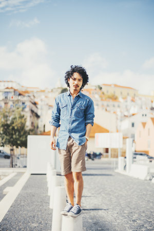 A young biracial man with curly black hair stands confidently on a concrete pillar in an urban outdoor setting. He wears a denim shirt, khaki shorts, and sneakers under a sunny blue skyの写真素材