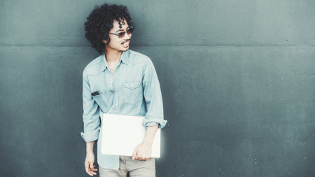 Young biracial man with curly hair wearing sunglasses and a casual denim shirt stands against a textured green wall, holding a closed laptop and looking to the sideの写真素材