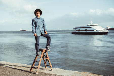 A young multiracial man with curly hair stands confidently on a wooden ladder by the waterfront, dressed in a denim shirt and jeans, with a ferry and ships in the background under a blue skyの写真素材