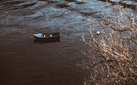 A small, empty wooden rowboat floating on a calm river with rippling dark water, surrounded by bare...