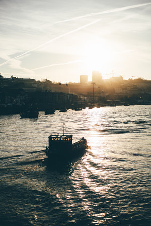 A scenic telephoto shot of riverscape at sunset featuring a boat sailing on calm waters, city silhouettes with cranes and cable cars in the background, and golden light reflectionsの写真素材