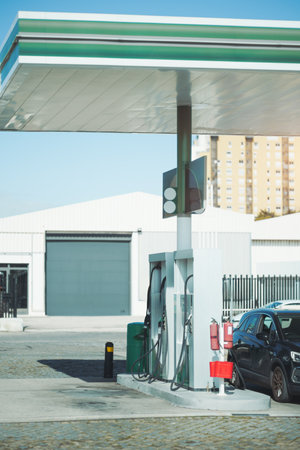 A modern gas station with fuel dispensers, fire extinguishers, and a parked car. The station has a green and white canopy, hoses hanging from pumps, and an industrial backdropの写真素材