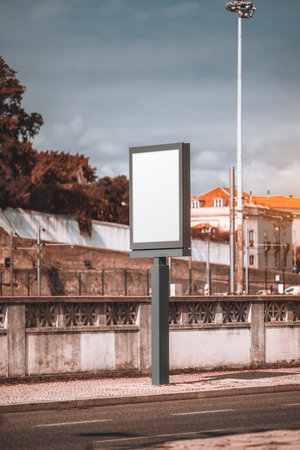 Blank vertical billboard mockup on a city street. Empty advertising panel with a modern frame on a sidewalk near a decorative concrete fence. Urban background with buildingsの写真素材