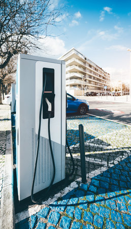 Electric vehicle charging station with a black charging cable and plug attached to a modern white charger unit in an urban setting. A blue car is parked nearby, with residential buildings behindの写真素材