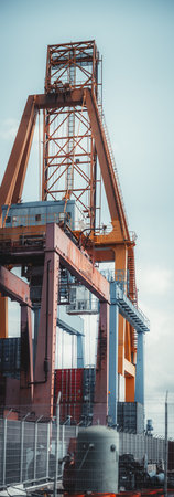 Telephoto vertorama of industrial port crane with rusted and weathered metal structure, steel framework, ladders, and platforms. Shipping containers and security fences in a commercial cargo terminalの写真素材