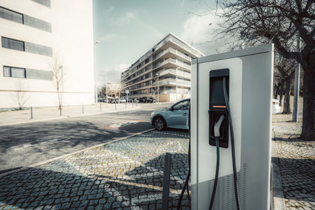 Wide-angle shot of a modern electric vehicle charging station on a cobblestone-paved urban street. A blue car is parked aloof. Trees, contemporary buildings, and soft sunlight create a cityscapeの写真素材