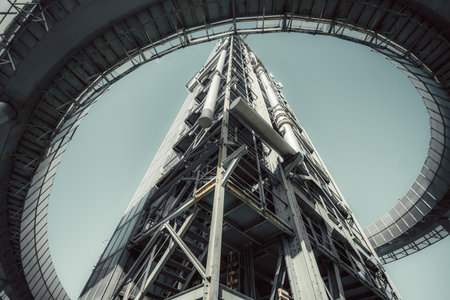 Low-angle view of a towering industrial metal structure with intricate pipes, beams, and a circular framework. The image captures a futuristic, sci-fi aesthetic with a muted skyの写真素材