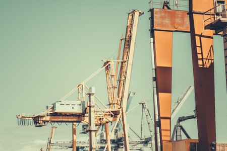Industrial port scene with large orange gantry cranes and container handling equipment under a clear sky. Steel structures, cables, and shadows create a dynamic composition; retro colours, telephotoの写真素材