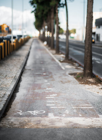 Weathered urban bike lane with faded white bicycle markings, cracked pavement, and scattered dirt. Parallel to a tree-lined road with parked cars and yellow safety barriersの写真素材