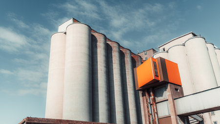 Industrial grain silos with a modern orange extension against a blue sky. Large cylindrical storage structures made of concrete, used for agriculture or food processing facilitiesの写真素材
