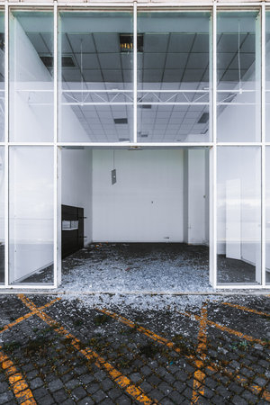 Abandoned commercial building ready for demolishing, with shattered glass at the entrance. Empty interior with white walls, broken ceiling panels, and scattered debris. Urban decay and vandalism sceneの写真素材
