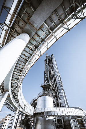 Wide-angle view of an industrial oil refinery structure with metal pipelines, a towering distillation column, and rusted steel framework under a clear blue sky, showcasing heavy industry architectureの写真素材