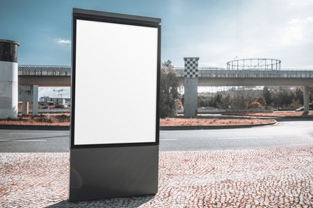 Outdoor advertising mockup with a blank vertical billboard in an urban setting. The empty ad space stands on a cobblestone sidewalk near a road, with industrial structures in the backgroundの写真素材