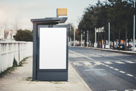 Modern bus stop with a blank vertical advertising billboard mock-up on a sidewalk near an empty city road. Urban environment with trees, road markings, and a pedestrian crossing near the templateの写真素材