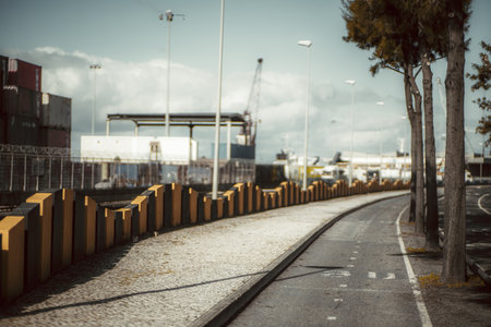 Urban landscape with bike path, sidewalk, industrial port, shipping containers, streetlights, fenced area, trees, curved road, and cloudy sky in the background. Telephoto shot; Lisbon; Portugalの写真素材