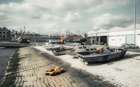 Old abandoned wooden boats decaying on a paved shore near industrial warehouses. A small yellow plastic toy boat lies on the ground. Urban background with cloudy skyの写真素材