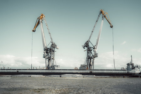 Two large industrial harbor cranes with yellow and gray arms stand symmetrically against a cloudy sky at a shipping dock. The foreground features a cobblestone pavementの写真素材