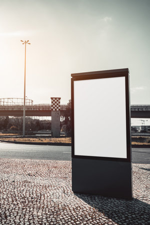 Outdoor urban mockup billboard with a blank white screen, set against a road, patterned pavement, and an industrial bridge structure. Ideal for advertising and branding designの写真素材