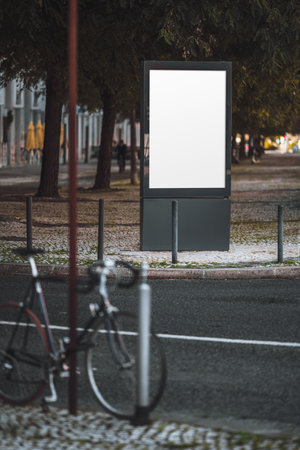 Urban mockup of a blank vertical advertising billboard on a city street sidewalk, surrounded by trees and blurred foreground bicycle. Ideal for showcasing poster design outdoors; vertical shotの写真素材