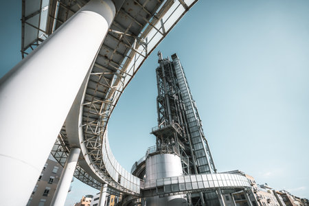 Low-angle view of modern industrial architecture featuring steel beams, curved elevated walkway, futuristic metallic structures, and buildings against clear blue sky background; wide-angle shotの写真素材