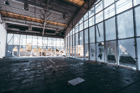 Interior of abandoned industrial building prepared for demolition with broken windows, shattered glass, damaged ceiling, debris on dirty floor, vandalism evidence, daylight sceneの写真素材