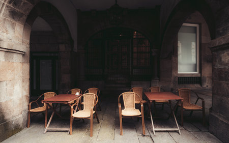 Empty outdoor cafe terrace under stone arches with wooden tables and wicker chairs, moody lighting, historic European architecture, cozy atmosphere; Santiago de Compostela, Spainの写真素材