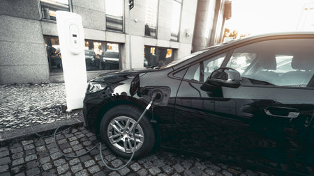 Modern black electric car charging at a white urban charging station on cobblestone street near a glass office building in the city, symbolizing sustainable mobility; wide-angle shot, Lisbon, Portugalの写真素材