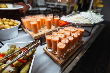 Close-up of a catering table with rows of small glasses filled with chilled orange gazpacho soup, pickles, green olives, and sliced cheese arranged in white dishes; wide-angle indoor shotの写真素材