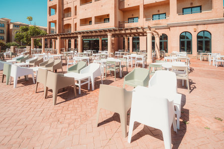 Outdoor hotel terrace with modern plastic chairs and tables arranged in rows on red brick paving, facing a Mediterranean-style building with arched windows and balconies; wide-angle shotの写真素材