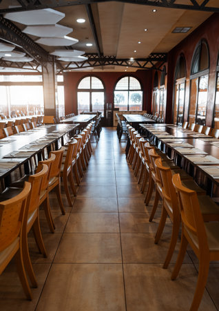 Elegant empty restaurant interior with long wooden tables and rows of matching chairs prepared for a group event or banquet, warm sunlight shining through arched windows; wide-angle vertical shotの写真素材