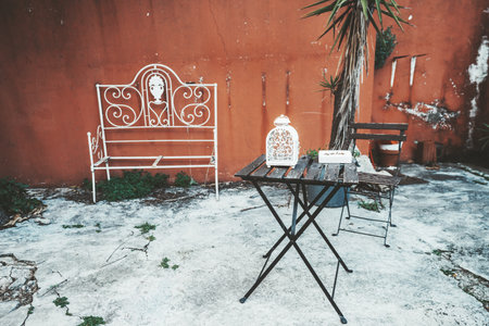 Outdoor patio scene with vintage metal furniture including a decorative white bench, a rustic table with a white lantern, a wooden chair, and a potted plant against a red wall; Lisbon, Portugalの写真素材