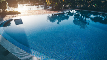 Tranquil early morning scene at a luxury resort swimming pool with calm blue water, tile details, tropical palm reflections, and sunlit lounge chairs in the background; Punta Umbria, Spainの写真素材