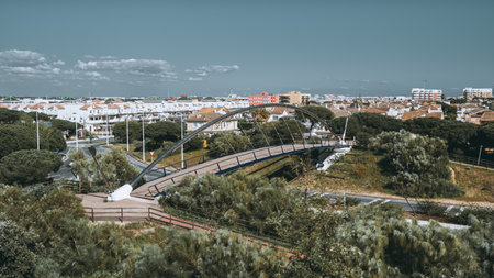 Modern pedestrian bridge with sleek metal arch crossing over green urban park, surrounded by white Mediterranean-style houses and pine trees in Punta Umbria, Andalusia, Spainの写真素材