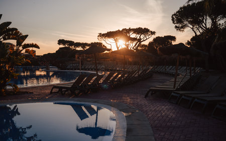 Sunset view of empty tropical resort pool area with rows of sun loungers, straw parasols, and calm water reflections surrounded by exotic trees, a peaceful vacation atmosphere; low-key wide-angle shotの写真素材