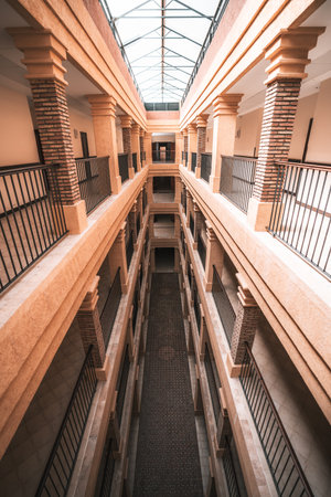 Bright interior view of a symmetrical multi-story corridor with brick columns, iron railings, and a glass ceiling allowing sunlight to illuminate the warm-toned architecture; wide-angle vertical shotの写真素材