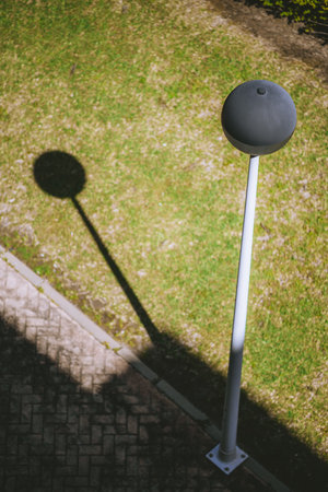 Modern street lamp casting a round, sharp shadow on green grass and pavement in bright sunlight, captured from above, creating a minimalist and geometric urban scene; telephoto shot, vertical formatの写真素材