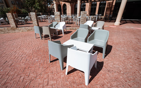 Modern outdoor patio with empty plastic chairs and white tables on red patterned tiles, sunlit terrace area of a restaurant or hotel, no people, sunny clear dayの写真素材