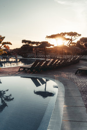 Serene resort poolside scene at sunset with empty lounge chairs, thatched parasols, and still water reflecting the soft light and silhouettes of trees, evoking relaxation and summer tranquilityの写真素材
