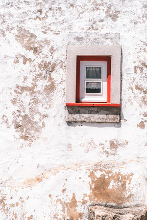 Small window with red trim set into a textured, weathered white wall, showing signs of age and peeling paint, with rustic charm and copy space ideal for text or design overlayの写真素材